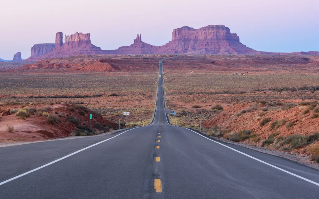 Lang, lige landevej mod de røde klippeformationer i Monument Valley under en lyserød himmel