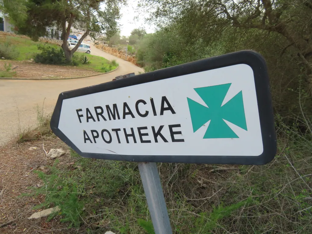 Vejskilt med teksten Farmacia og Apotheke og et grønt apotekerkors ved en grusvej på Mallorca