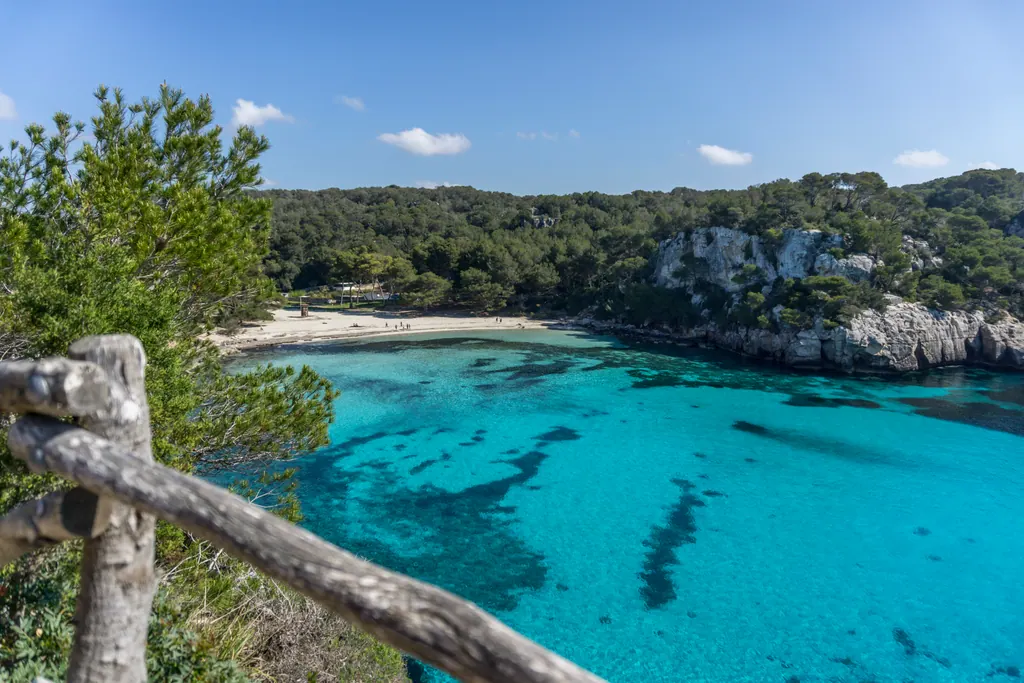 Turkisblåt vand og hvid sandstrand i bugten Cala Macarella på Menorca, omgivet af fyrreskov og kalkklipper