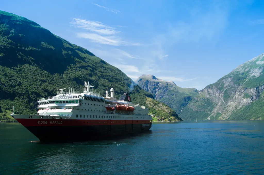 Hurtigrutens skib MS Kong Harald sejler i Geirangerfjorden mellem grønne fjelde under en blå sommerhimmel